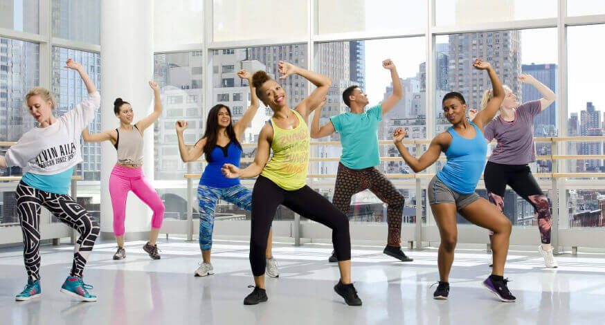 Turma em aula de dança numa sala ampla e iluminada, todos com os braços erguidos.