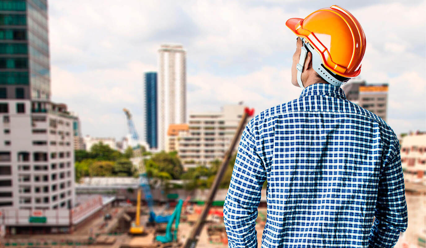 Engenheiro de capacete laranja, de costas, observando prédios e guindastes de um canteiro de obras.
