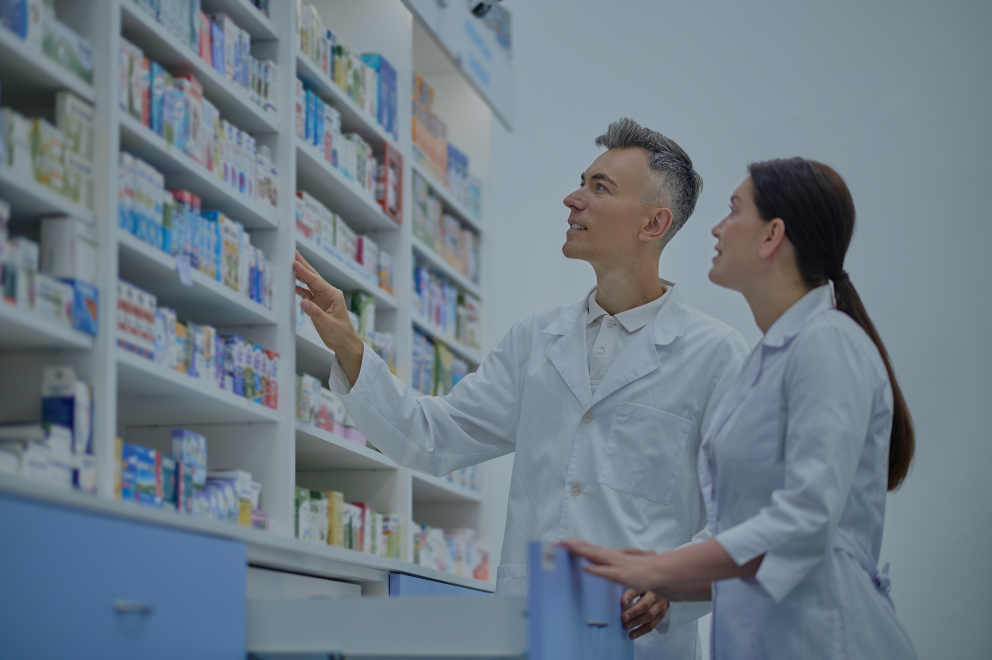 Dois farmacêuticos de jaleco branco conferindo medicamentos nas prateleiras de uma farmácia.