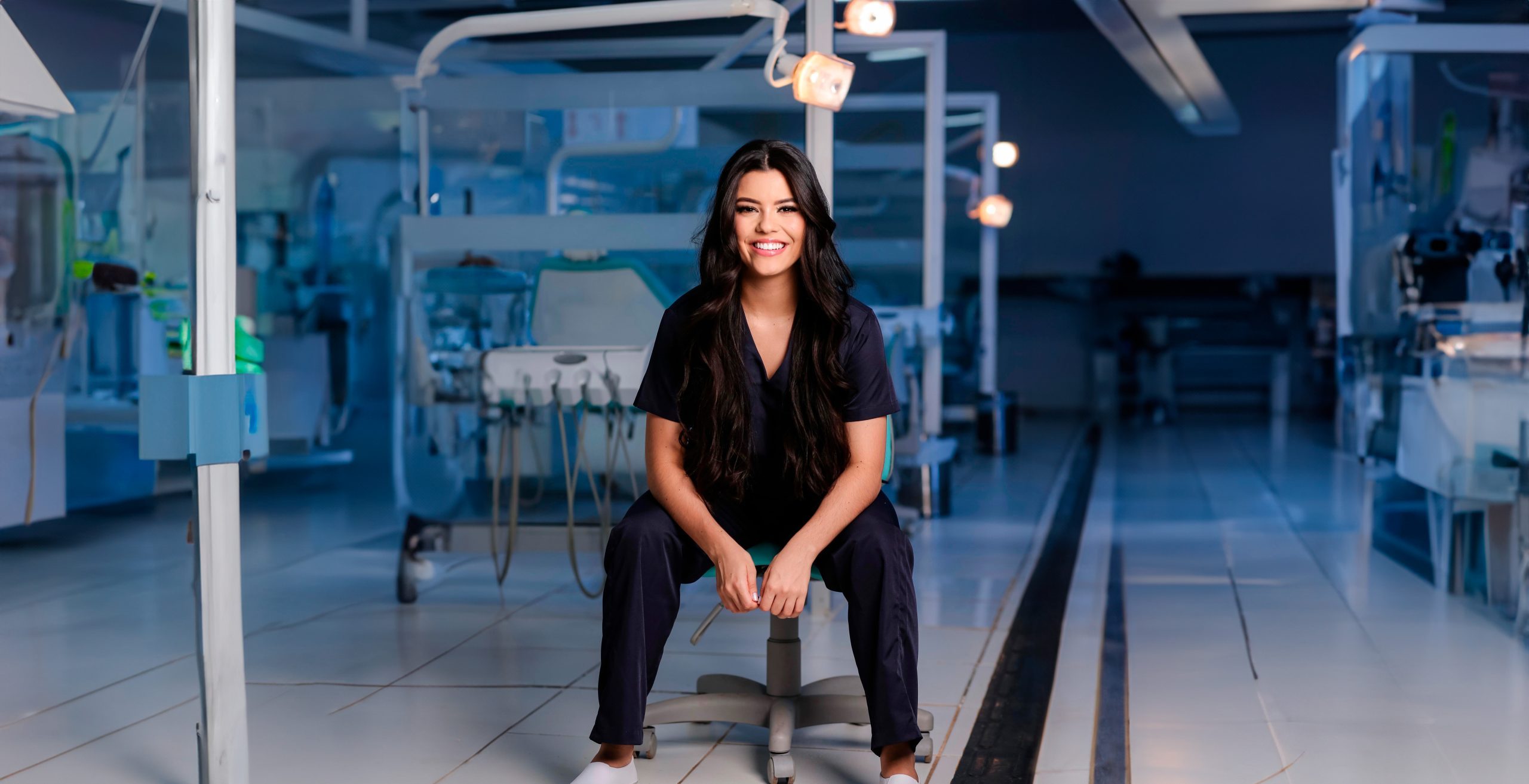 Estudante de Odontologia sorrindo, sentada na clínica odontológica da FAVENORTE, com cadeiras e refletores ao fundo.